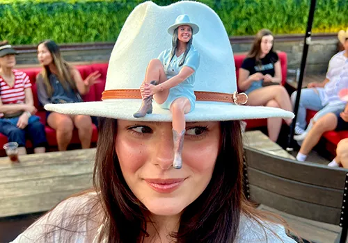 Miniature 3D-printed replica of a woman in a cowgirl hat, perched on the hat brim of the real person who was scanned